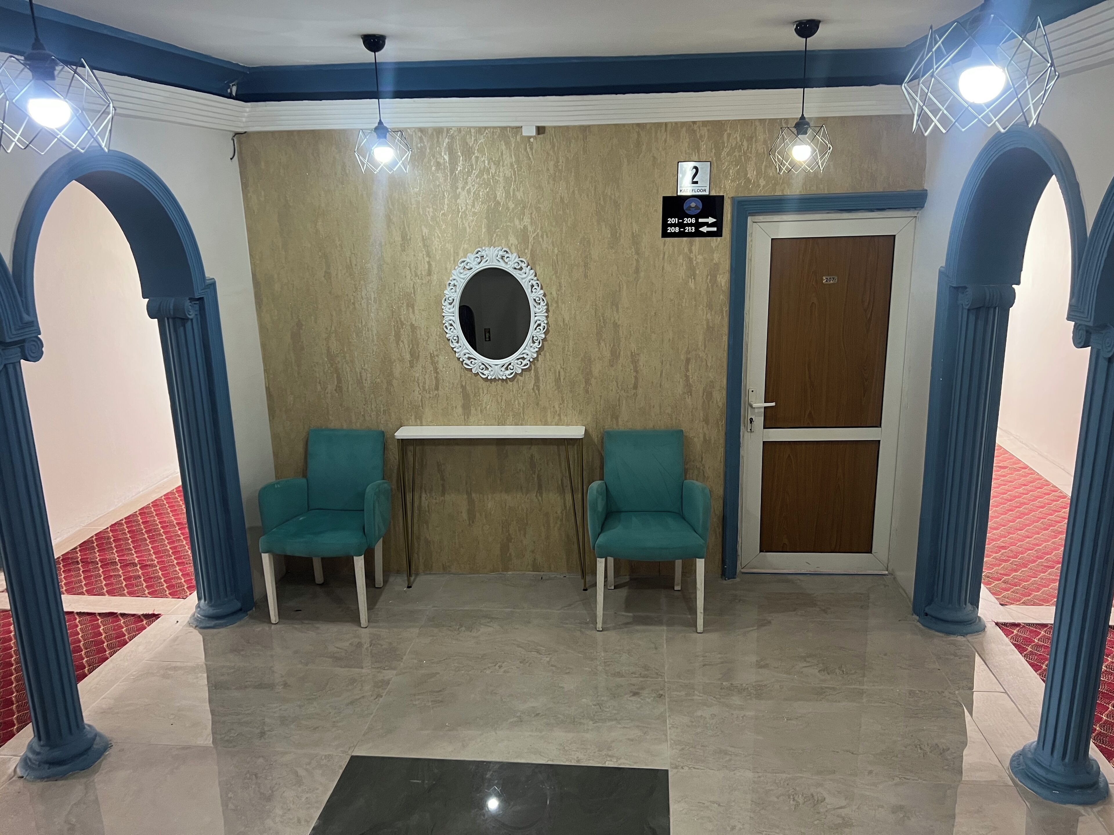 Un vestíbulo con dos sillas color turquesa, un espejo, un cartel con los números de las habitaciones y una puerta.