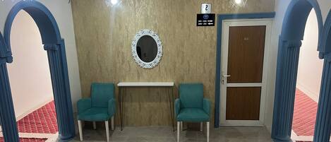 Un vestíbulo con dos sillas color turquesa, un espejo, un cartel con los números de las habitaciones y una puerta.