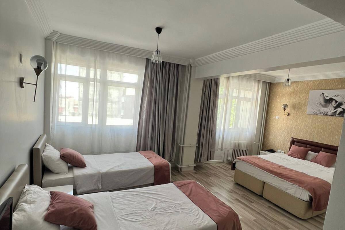 A hotel room with three beds, a large window with curtains, a wooden floor, and a wall-mounted light fixture.