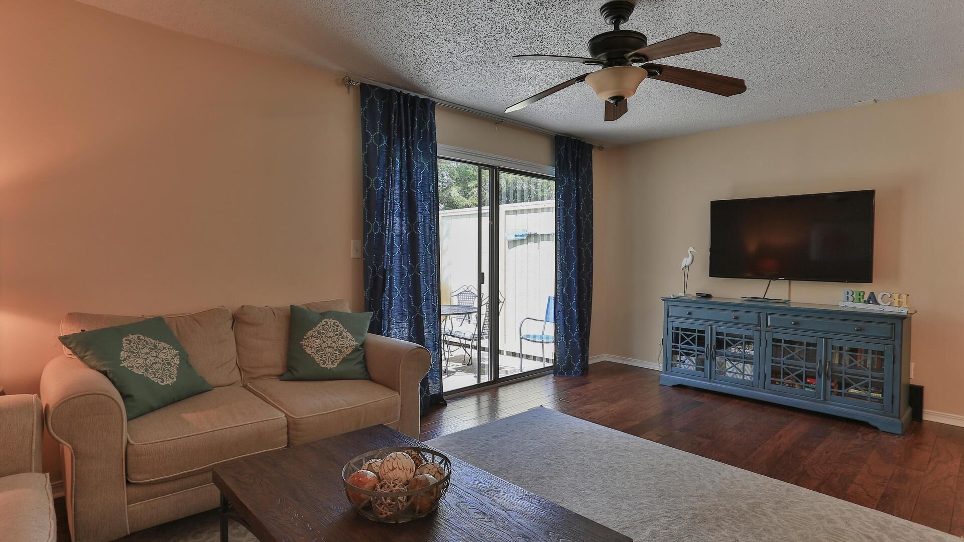 A living room with a beige sofa, a coffee table with decorative items, a ceiling fan, a TV on a blue cabinet, and a sliding glass door to the outside.