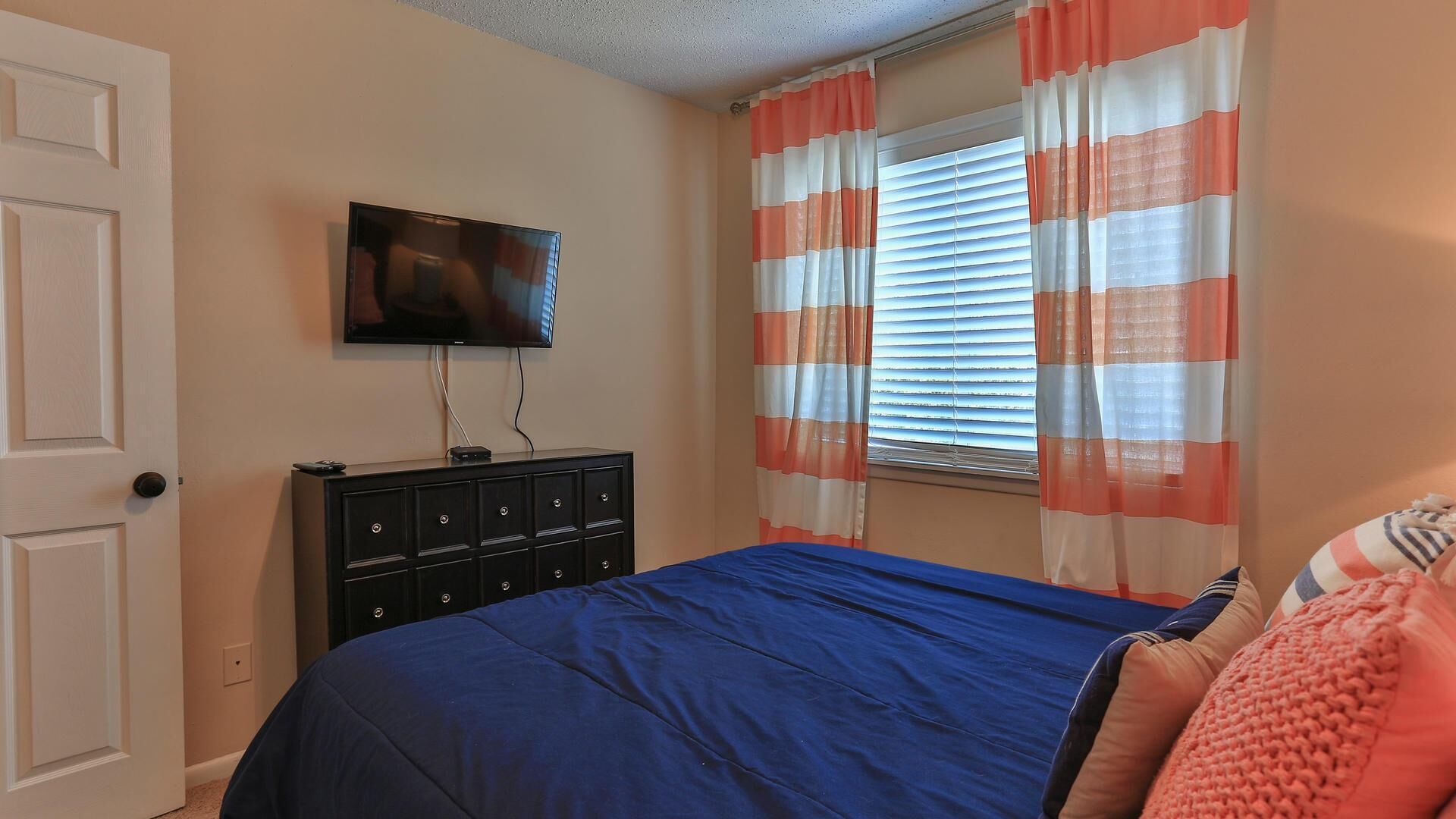 A bedroom with a bed, a dresser, a television, and a window with striped curtains.
