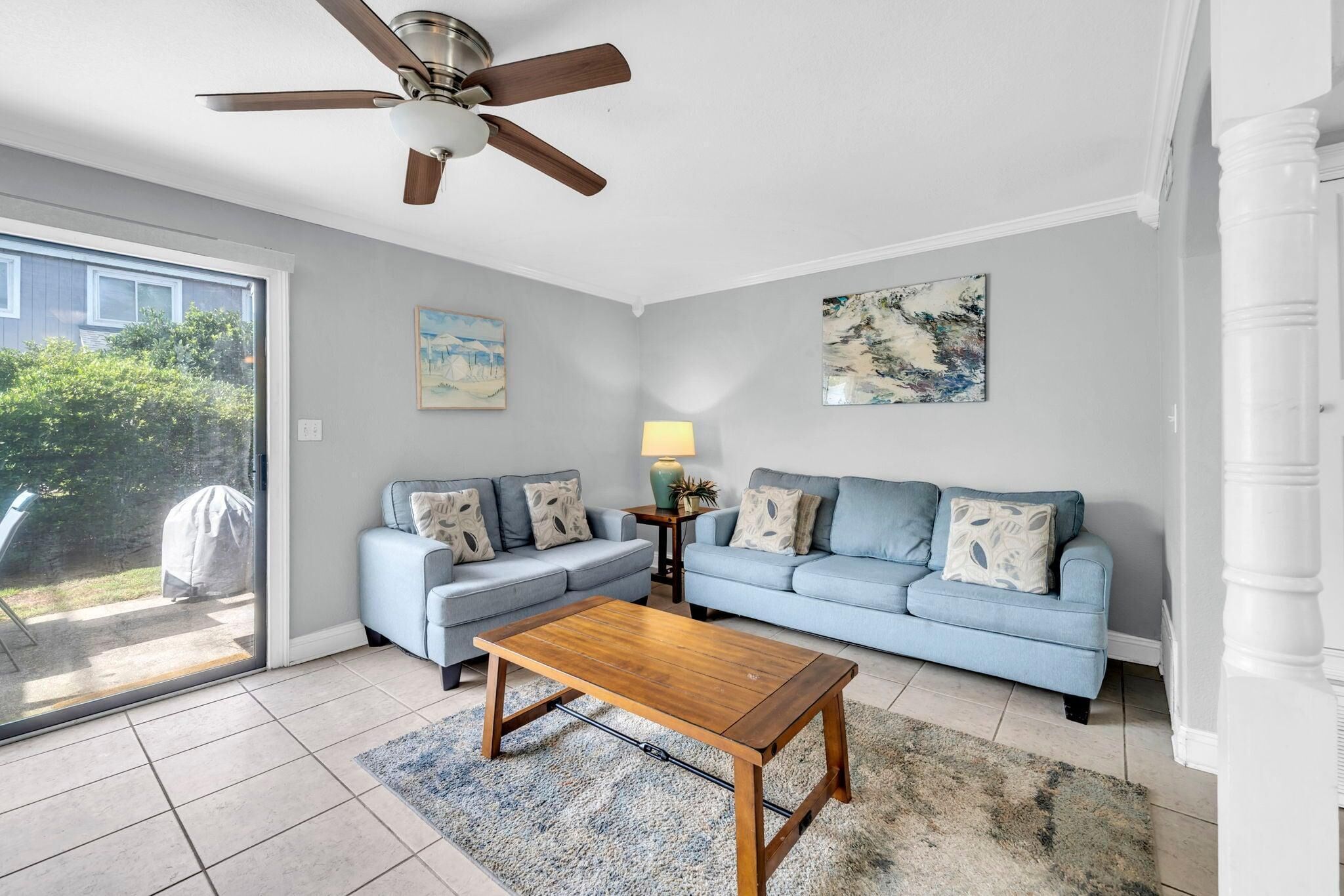 A living room with a ceiling fan, two light blue sofas, a wooden coffee table, and a glass door leading outside.