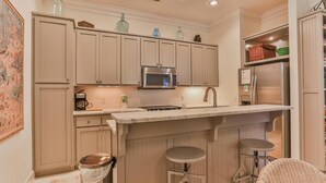 A kitchen with light wood cabinets, a stainless steel microwave, a sink, and a bar area with stools. - Conch Cottage (Santa Rosa Beach)