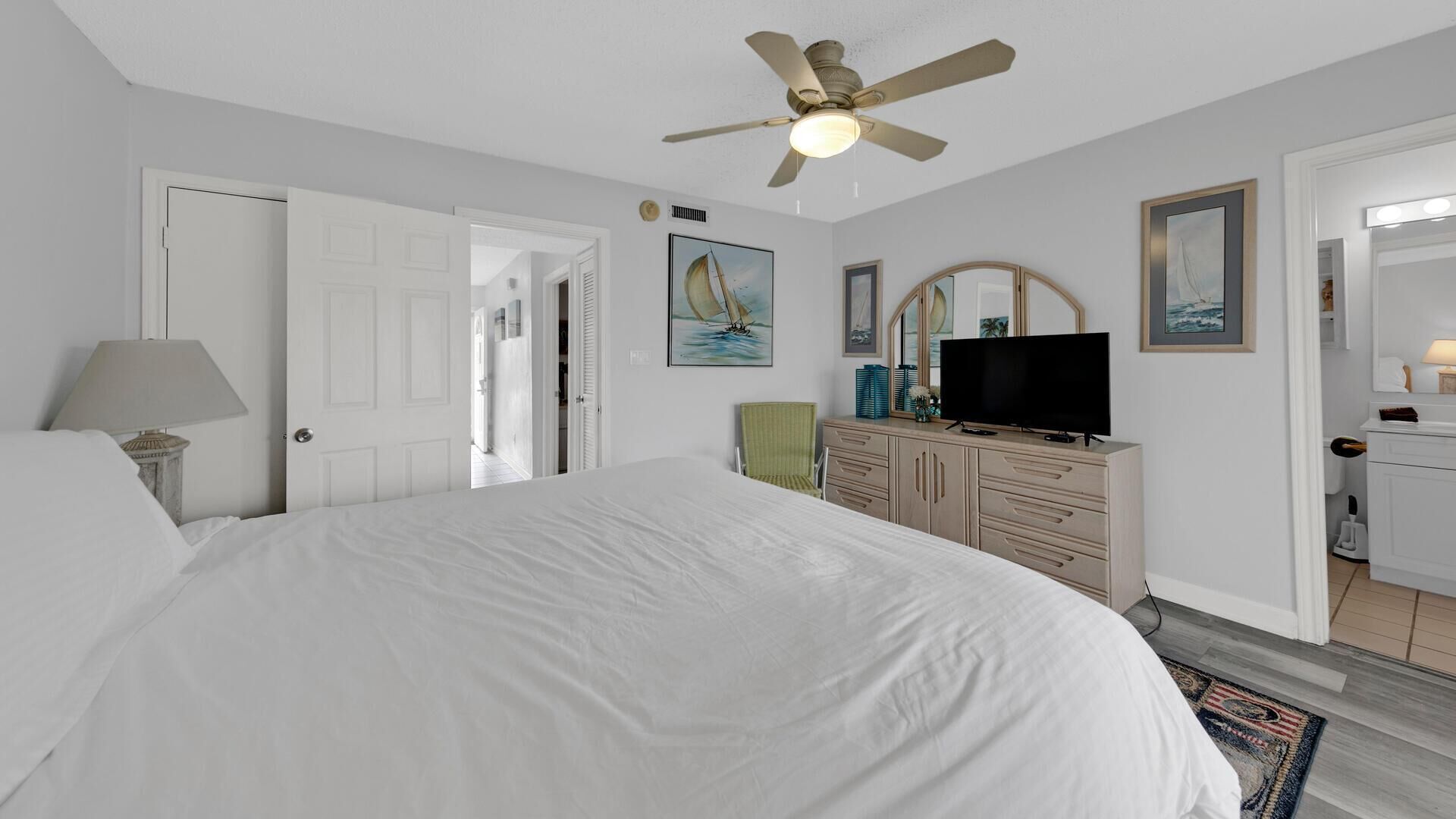 Une chambre à coucher avec un grand lit, un ventilateur de plafond, une télévision, une commode et une porte donnant sur une autre pièce.