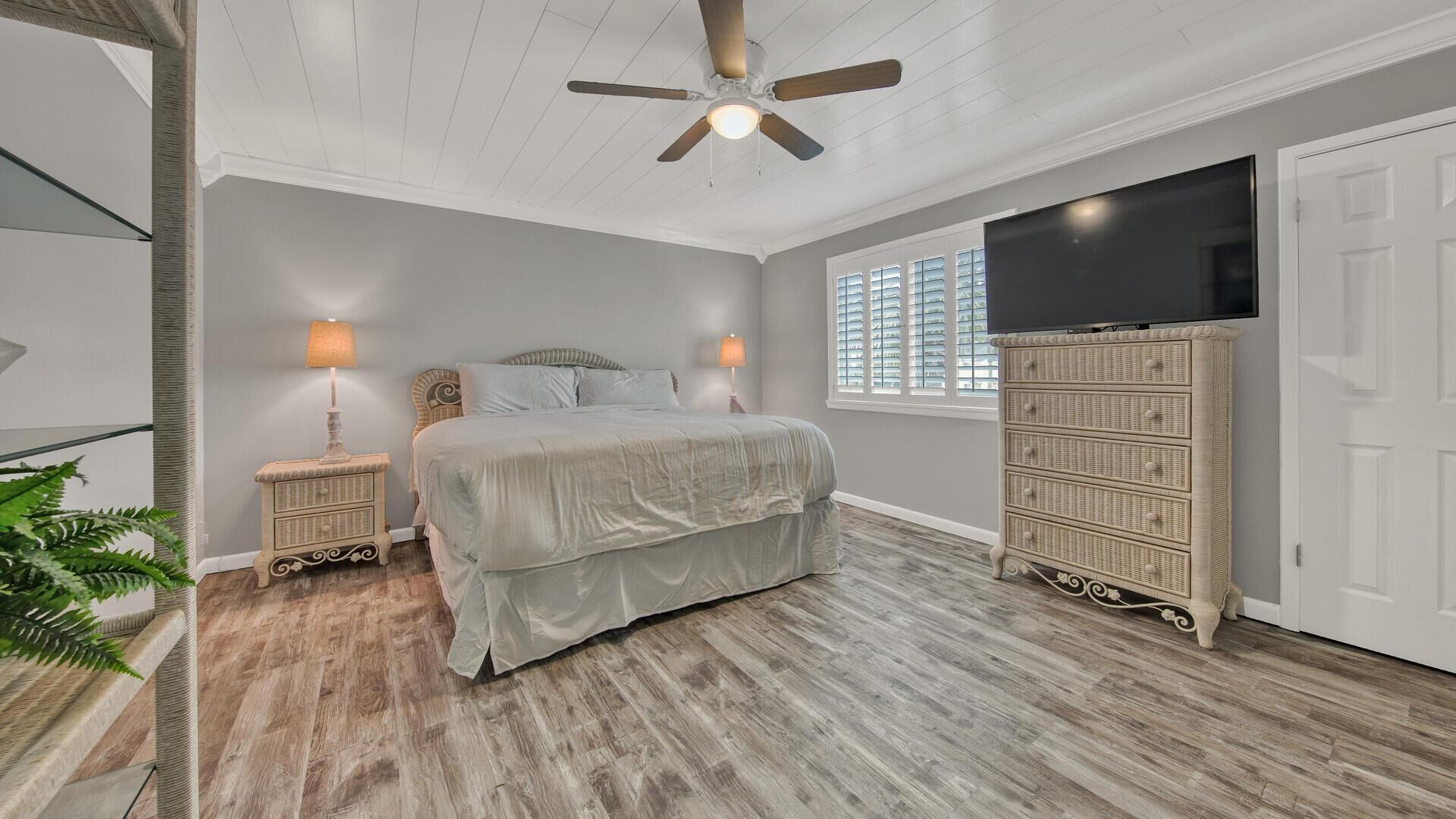 A bedroom with a bed, two bedside lamps, a ceiling fan, a television, and a wicker dresser.