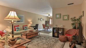 A living room with a sofa, coffee table, and a dining area with chairs and a table. There is a ceiling fan, a bookshelf, and a plant. - Southbay by the Gulf 73 (Destin)