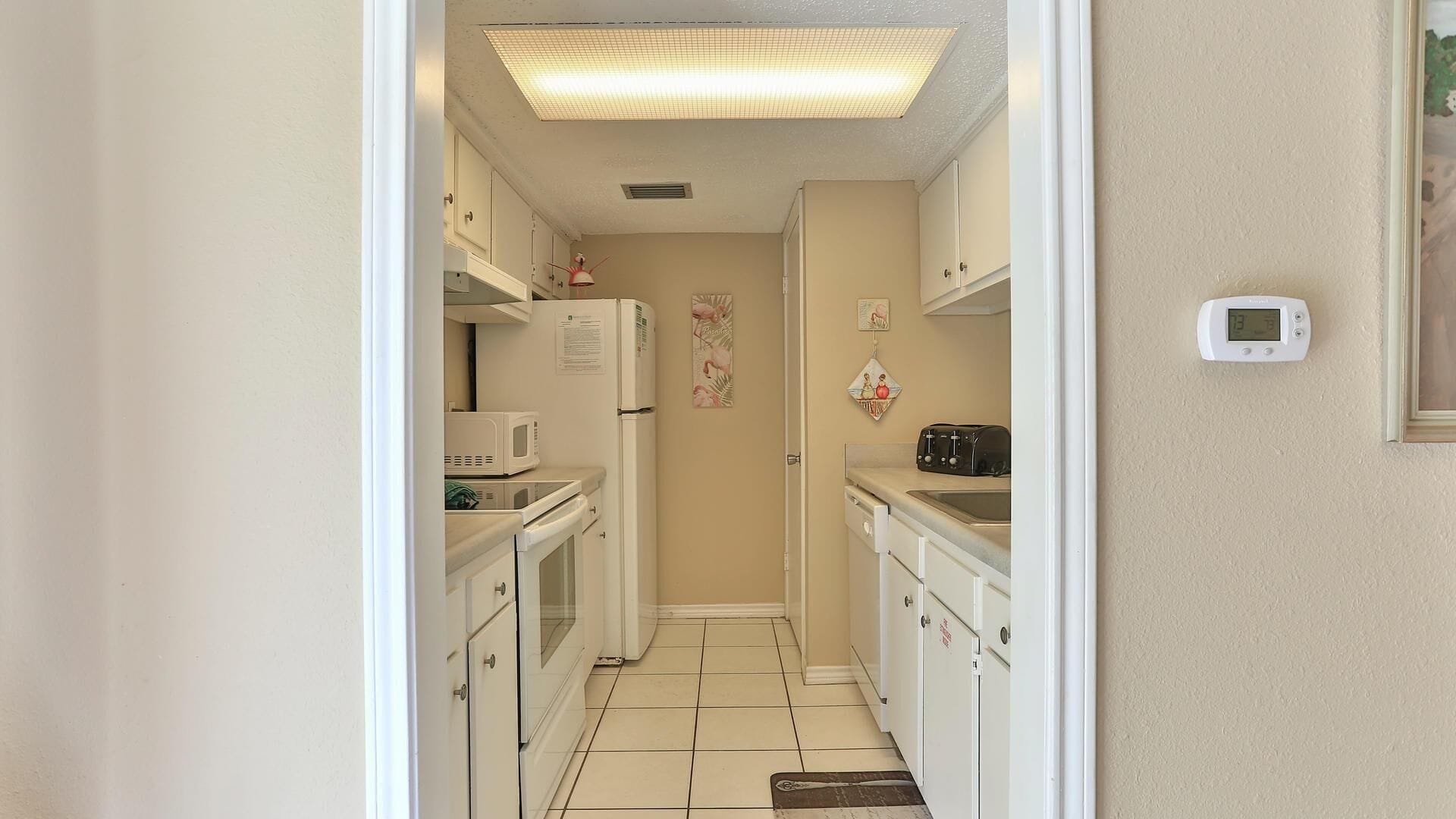 A narrow kitchen with white cabinets, a microwave, a toaster, and a refrigerator. There is a wall-mounted thermostat and a small framed picture on the wall.