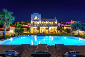 Una casa grande con piscina, sillas de descanso y un jardín iluminado durante la noche.