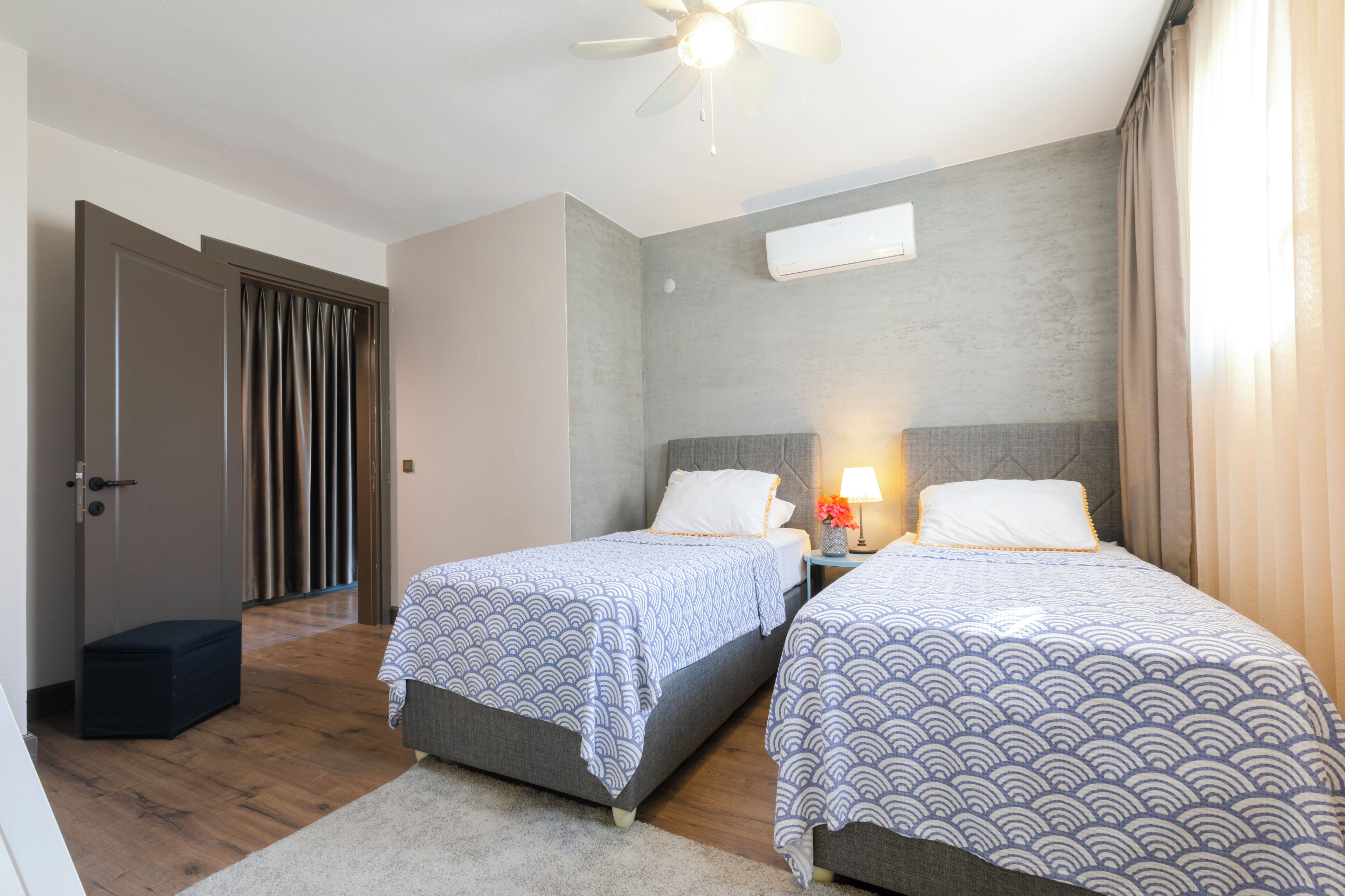 A hotel room with two single beds, a ceiling fan, an air conditioning unit, and a small bedside table with a lamp.