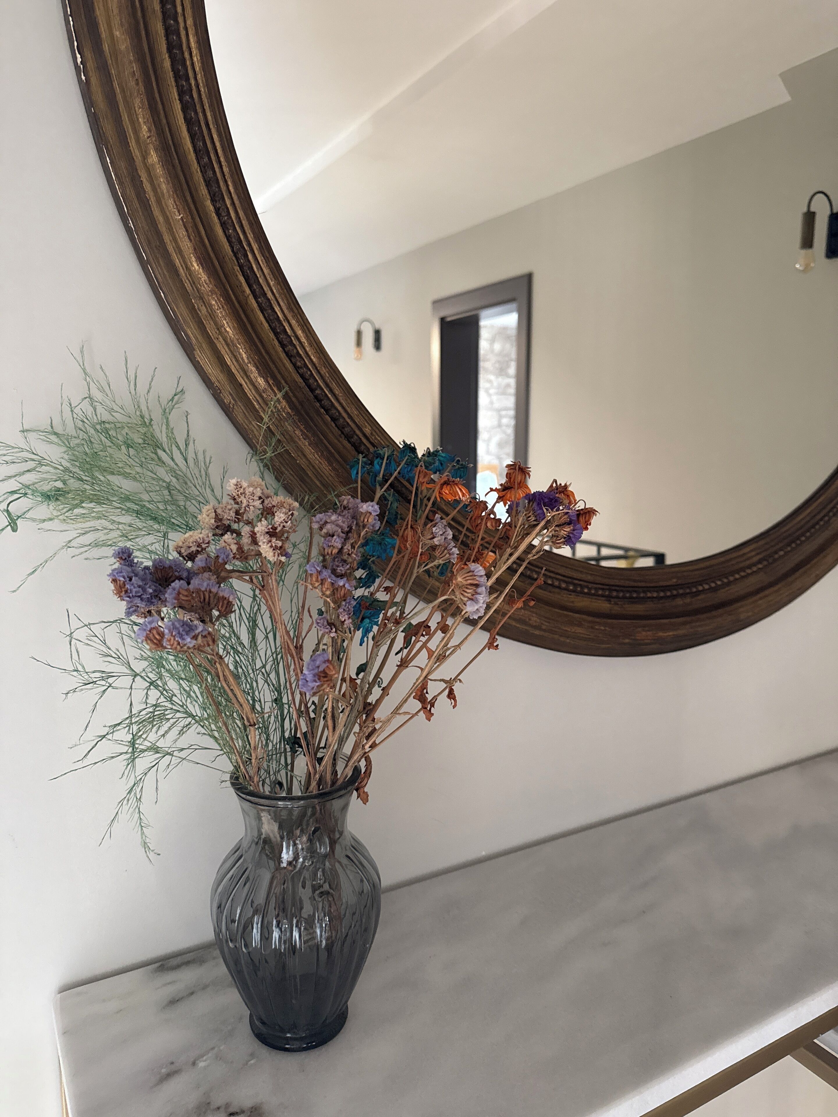 Un vase avec des fleurs séchées sur un comptoir en marbre, un grand miroir au mur et une porte à l’arrière-plan.