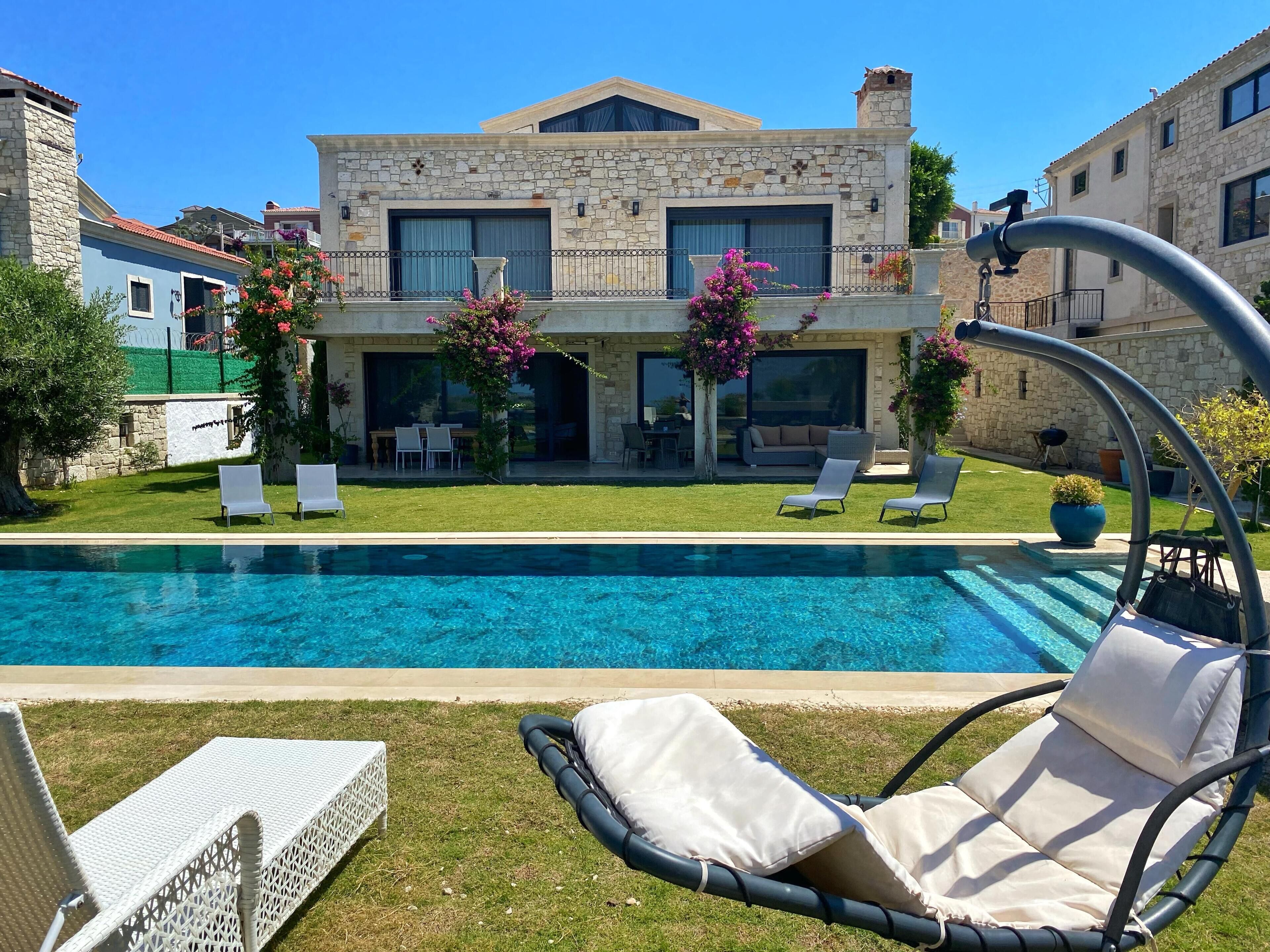 A poolside view of a stone house with a swimming pool, outdoor seating, and a well-maintained lawn.