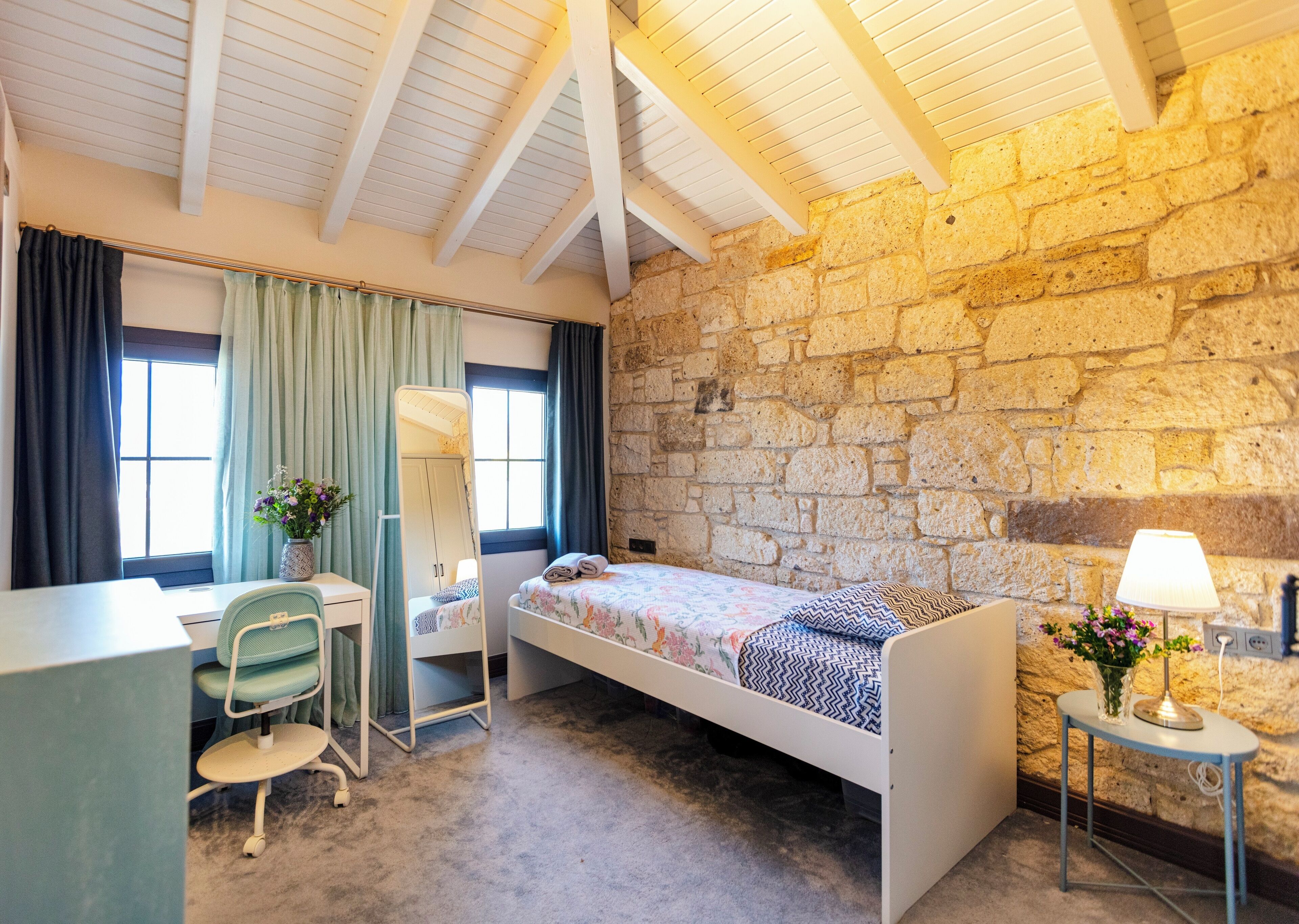 A bedroom with a bed, a desk, a chair, a mirror, and a stone wall.