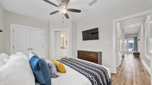 A bedroom with a bed, a ceiling fan, a TV, and a hallway leading to a balcony. - Hidden Lake Condos 113 (Santa Rosa Beach)