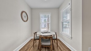 A dining area with a round table and four chairs, a clock on the wall, and a window with blinds. - Hidden Lake Condos 113 (Santa Rosa Beach)