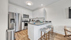 A modern kitchen with white cabinets, stainless steel appliances, a granite countertop, and a wooden bar stool. - Hidden Lake Condos 113 (Santa Rosa Beach)
