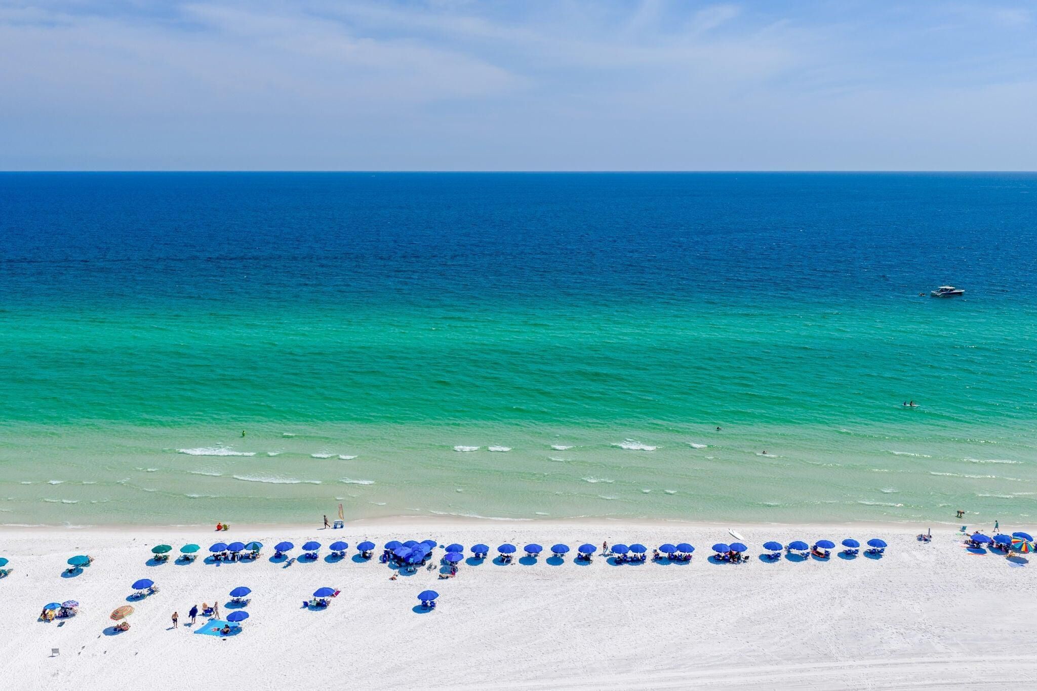 Une plage de sable blanc bordée d’une eau turquoise et cristalline. Des parasols bleus et des chaises longues sont disposés le long du rivage.
