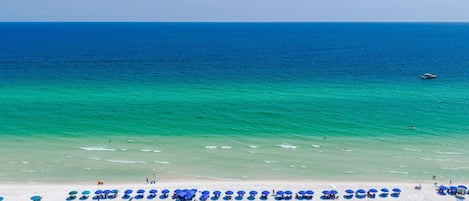 A beach with white sand and clear turquoise water. There are blue umbrellas and lounge chairs along the shore.