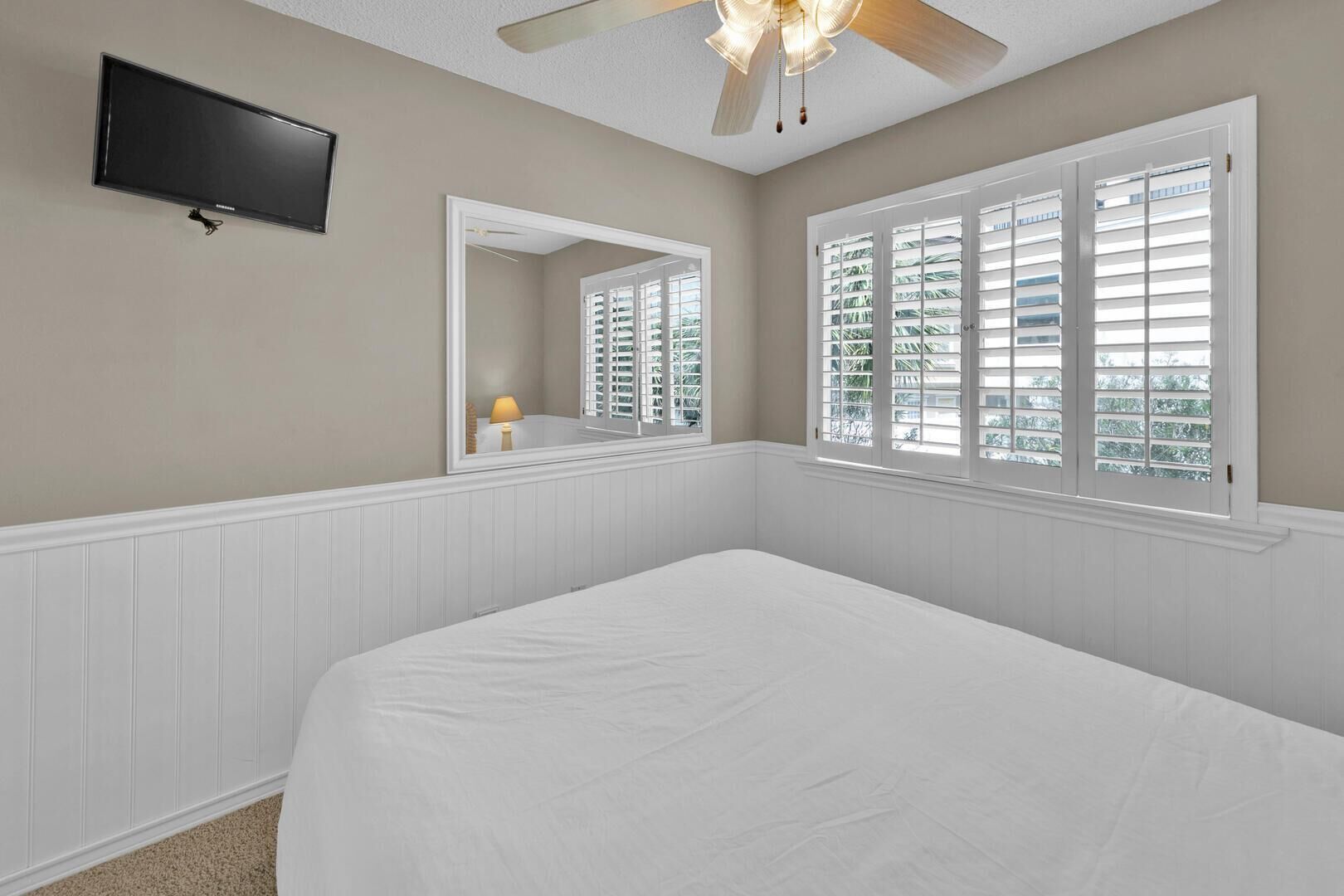A bedroom with a bed, a ceiling fan, a TV mounted on the wall, and windows with shutters.