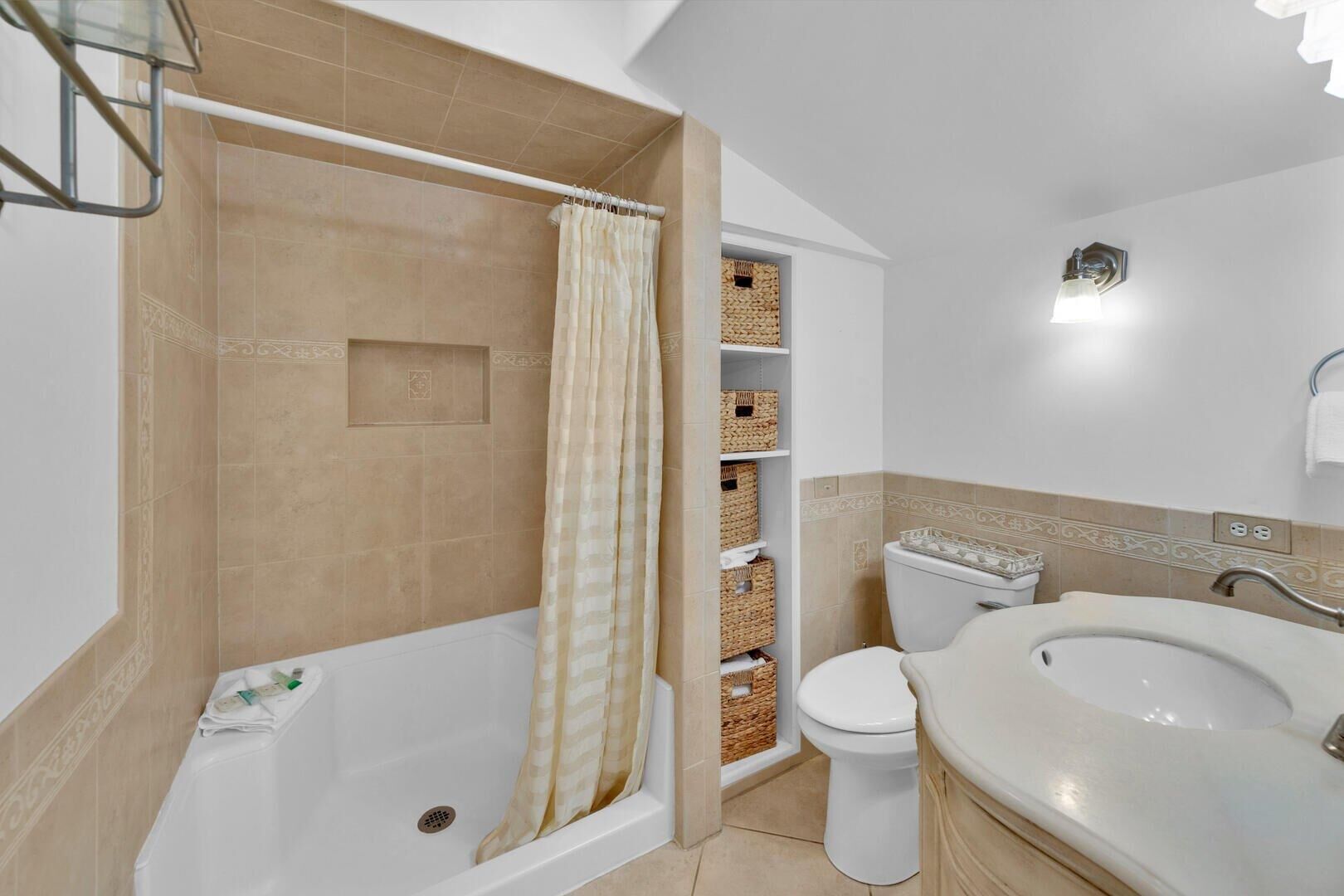 Een badkamer met bad, toilet en wastafel. Er staan rieten manden op planken en er hangt een douchegordijn.