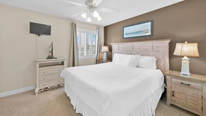 A bedroom with a large bed, a ceiling fan, a TV mounted on the wall, and a window with shutters.