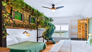 Une chambre avec un lit vert, un ventilateur de plafond, une fenêtre donnant sur un plan d’eau et un mur d’escalade.
