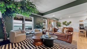 Un salon moderne avec un canapé, des fauteuils et une table basse. On y trouve également des plantes et une grande fenêtre.
