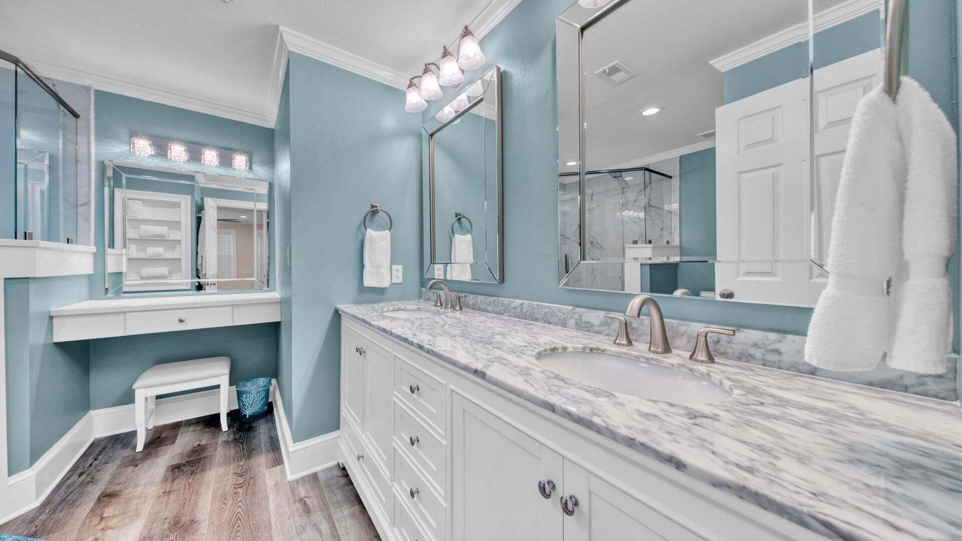 Une salle de bain avec un lavabo en marbre, des meubles blancs, une coiffeuse, un banc et un grand miroir avec éclairage.