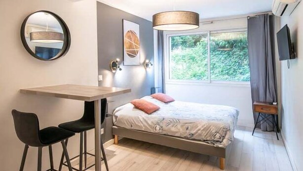A bedroom with a bed, a desk, a chair, a round mirror, a window with curtains, and a TV on the wall. - Central Part-Dieu - Apartment 2 - A/C available (Lyon)