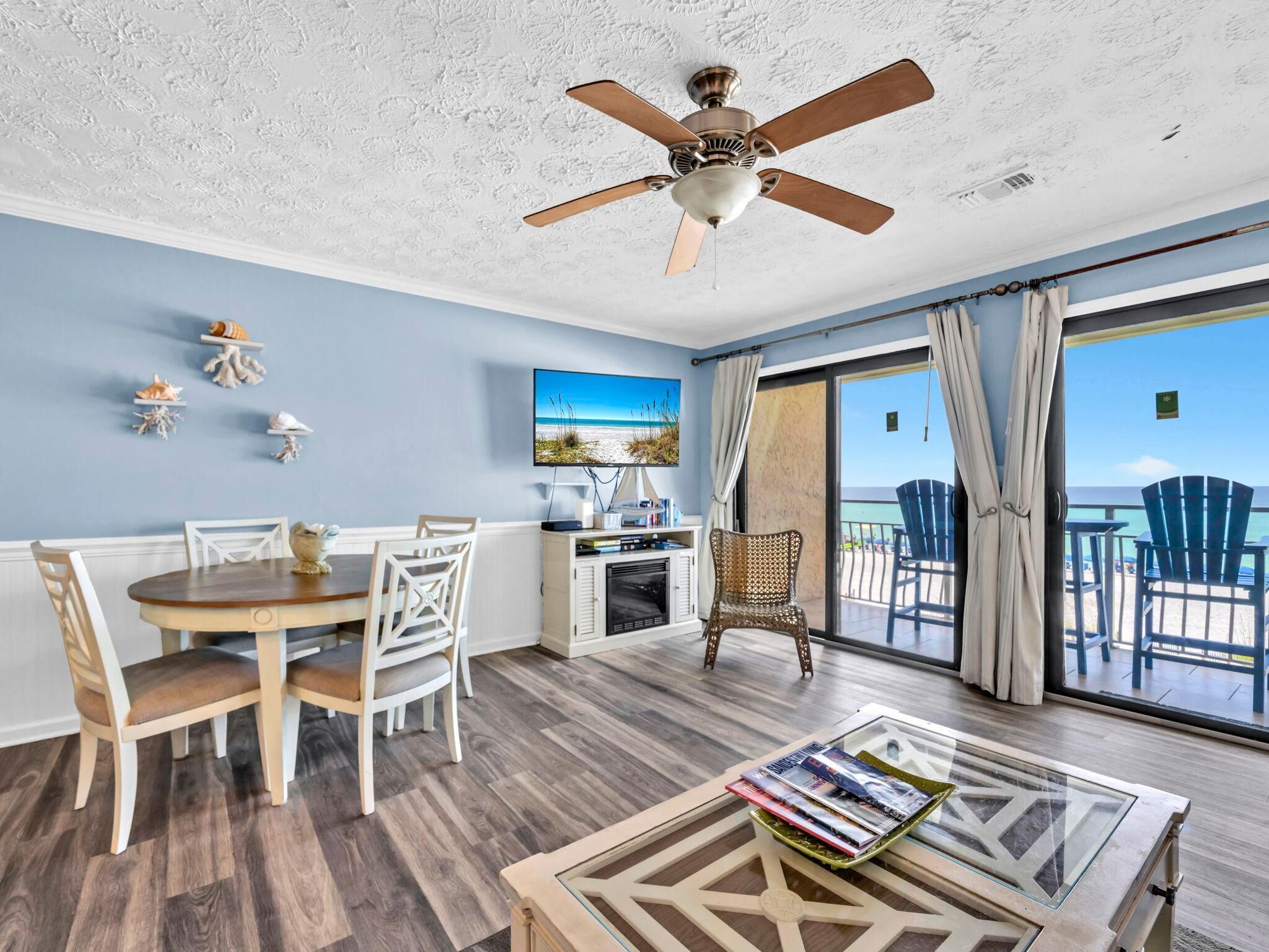 A room with a dining table, chairs, a TV, a ceiling fan, and a view of the beach through a large window.
