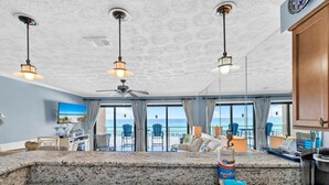 A spacious living area with a large window overlooking the ocean, a ceiling fan, and a kitchen counter with a coffee maker and baby formula. - Crystal Villas 6A (Destin)