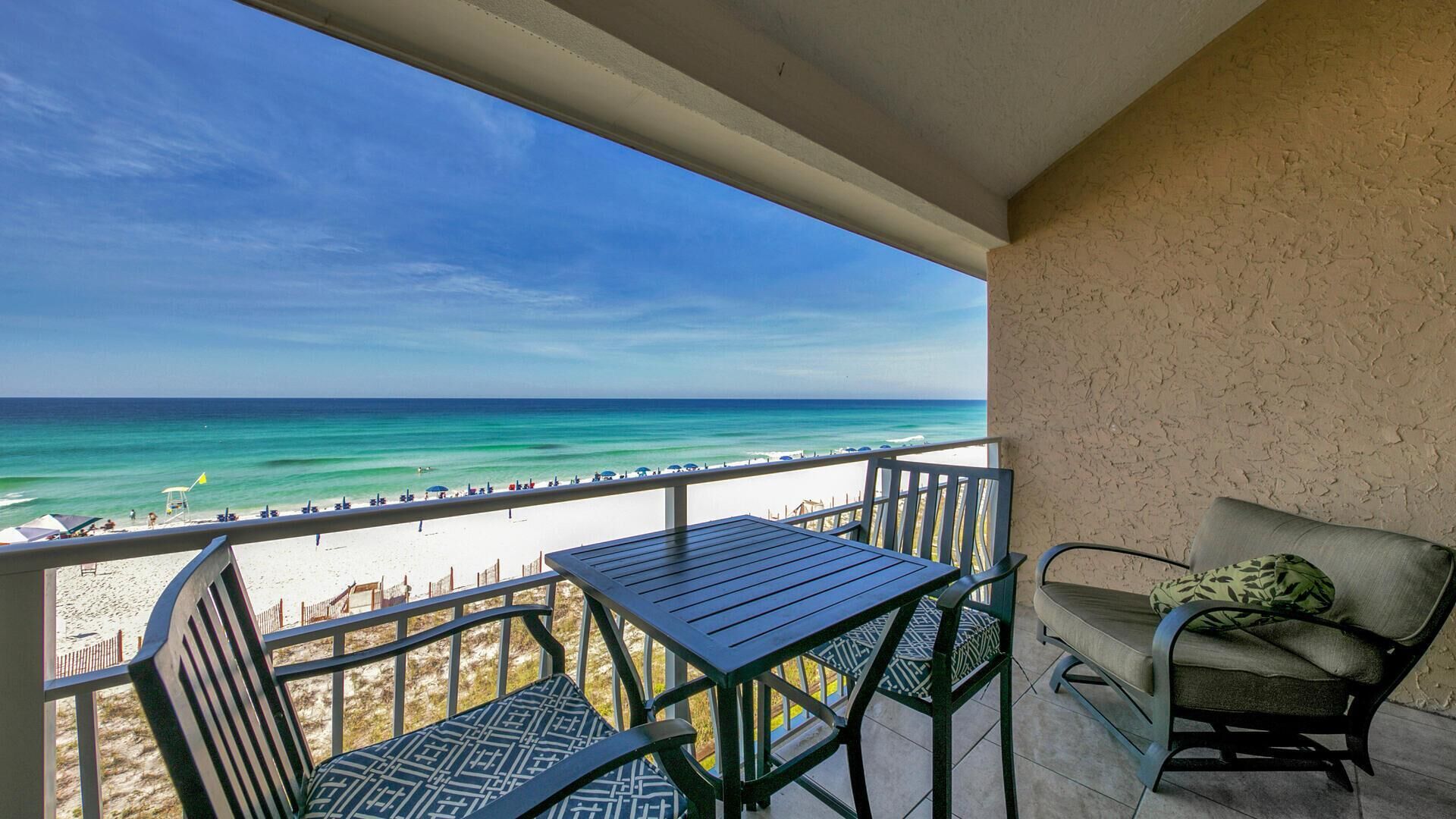 A balcony with a view of the beach, a table, and two chairs.