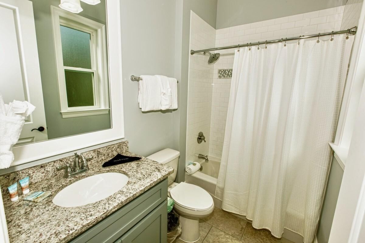 Une salle de bain avec un lavabo sur un plan de travail en granit, des toilettes, une douche avec un rideau et un miroir placé au-dessus d’une fenêtre.