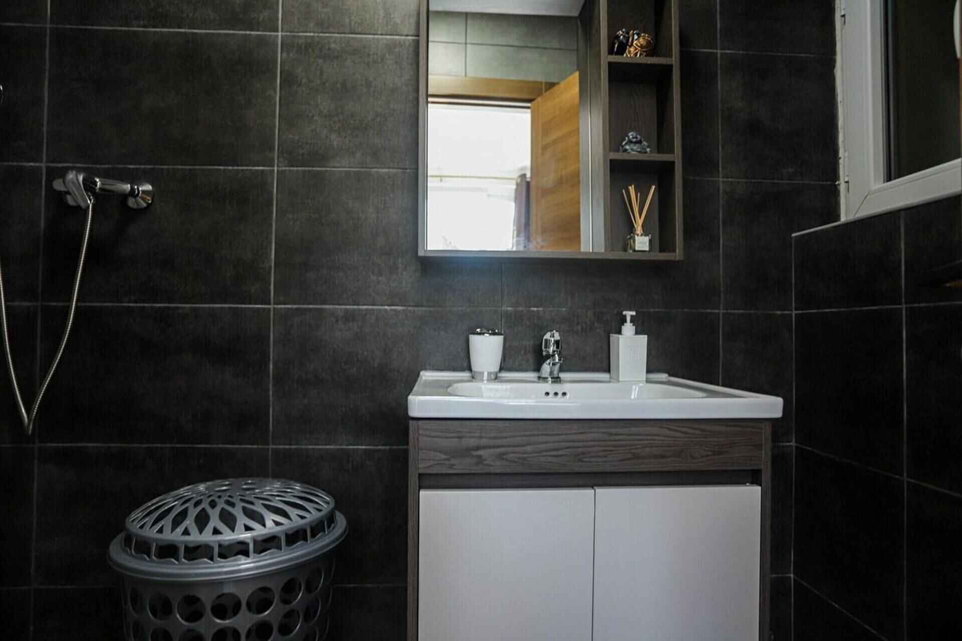 Un baño moderno con baldosas oscuras, un lavabo blanco, un espejo y una ducha.