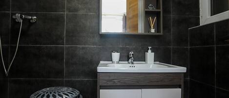 Un baño moderno con baldosas oscuras, un lavabo blanco, un espejo y una ducha.