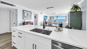 A modern kitchen with white cabinetry, a large island with a sink, and a view of the ocean through large windows. - Hidden Dunes Gulfside 206 (Miramar Beach)