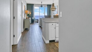 A narrow hallway with wooden flooring leading to a kitchen area with white cabinets and a dining area with a table and chairs. - Leeward Key 1103 (Miramar Beach)