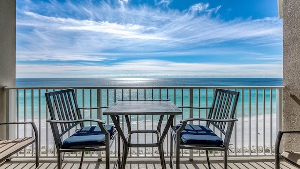 Ein Balkon mit Tisch und vier Stühlen, mit Blick auf einen Strand und klares türkisfarbenes Wasser unter blauem Himmel.