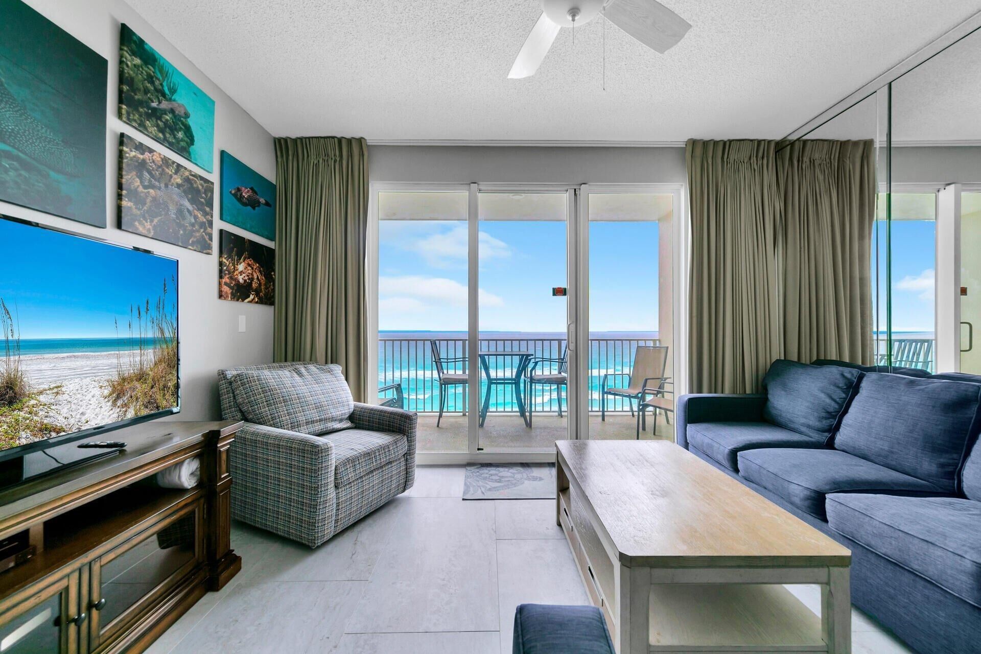 A living room with a sofa, armchair, and a coffee table. There is a TV mounted on the wall and a balcony with a view of the ocean.