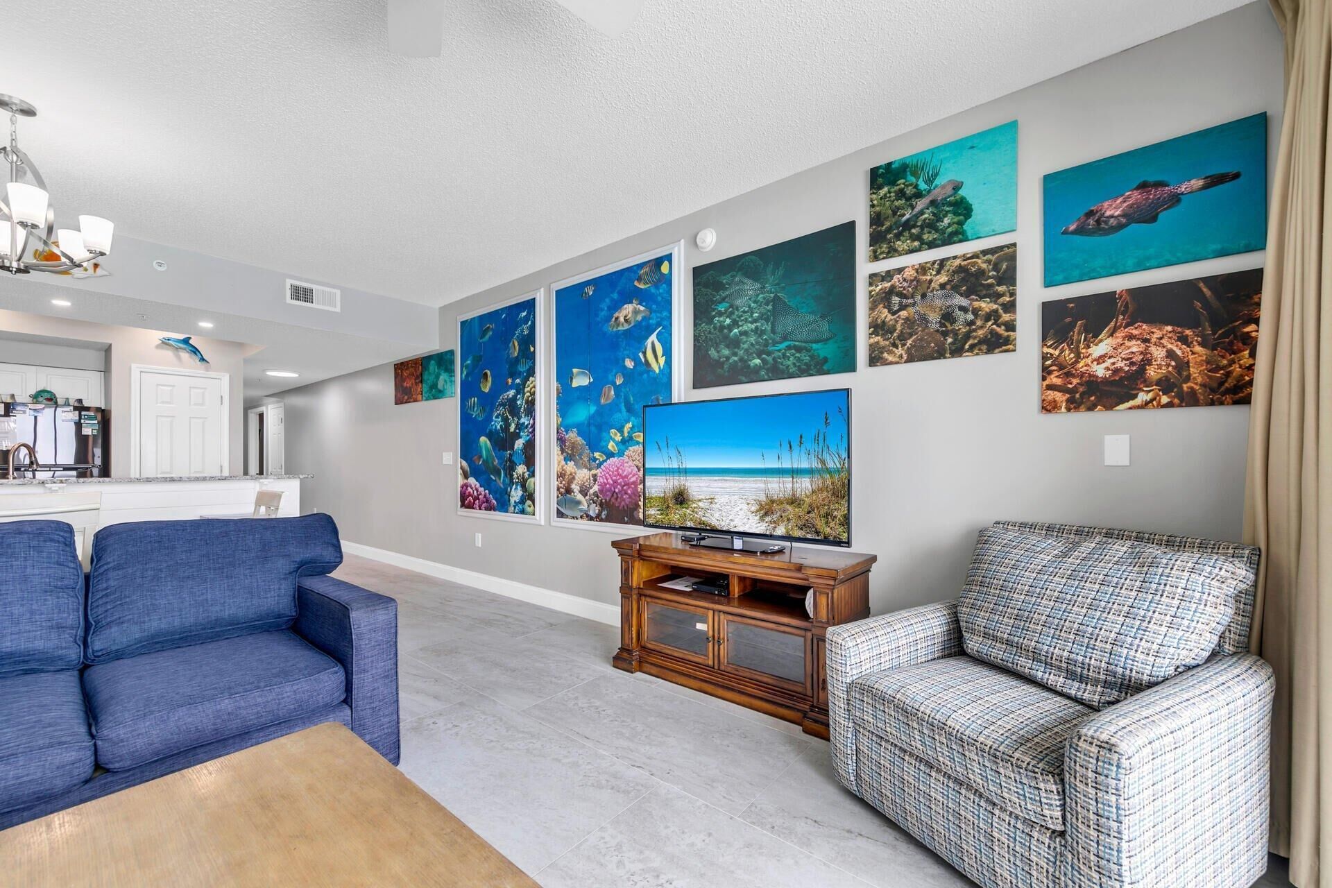 Un salón con un sofá azul, un sillón estampado, un mueble de televisión de madera y un televisor grande montado en la pared.