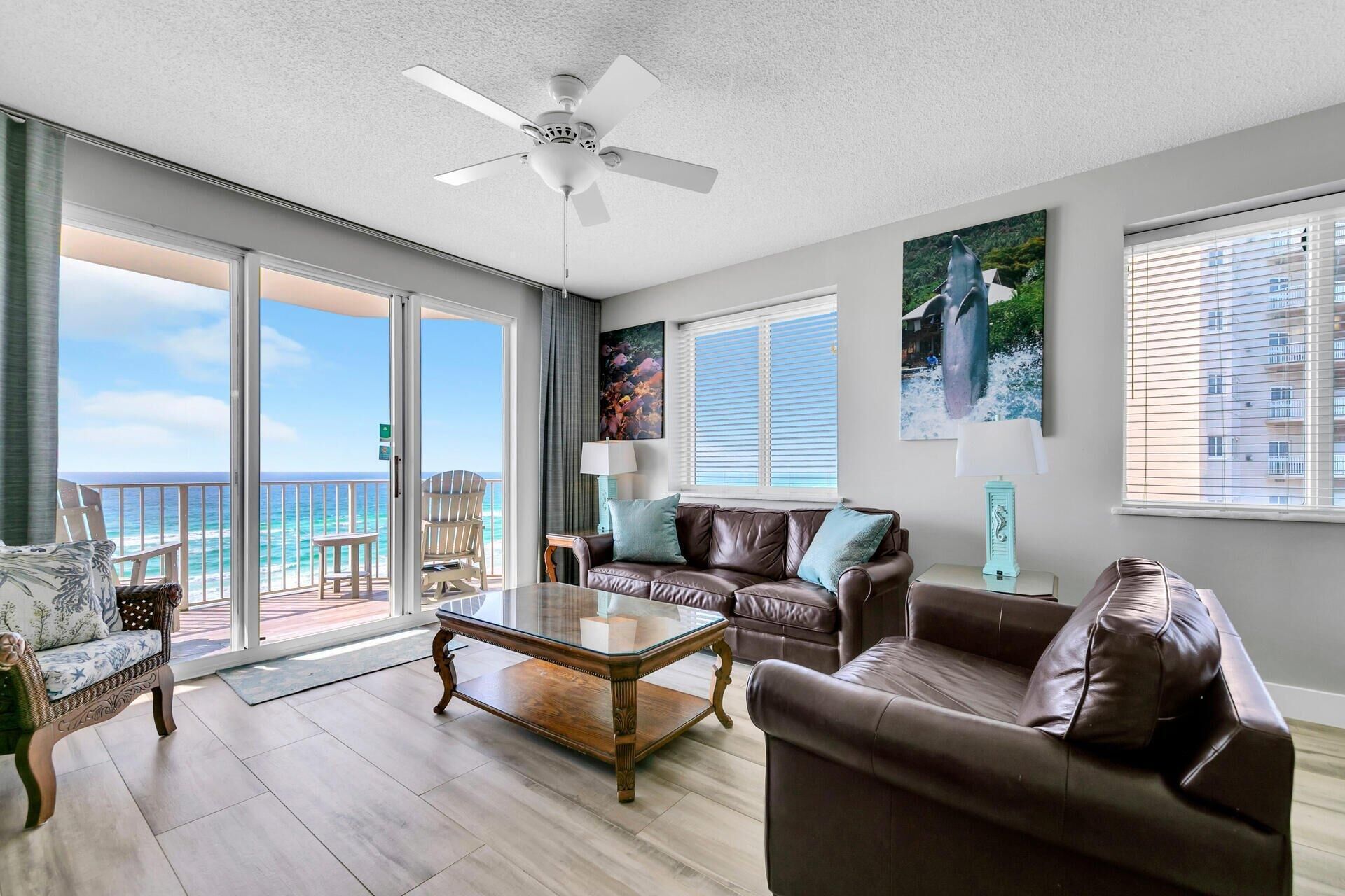 A spacious living room with a glass coffee table, leather sofas, and a ceiling fan. A balcony with a view of the ocean is visible through large windows.