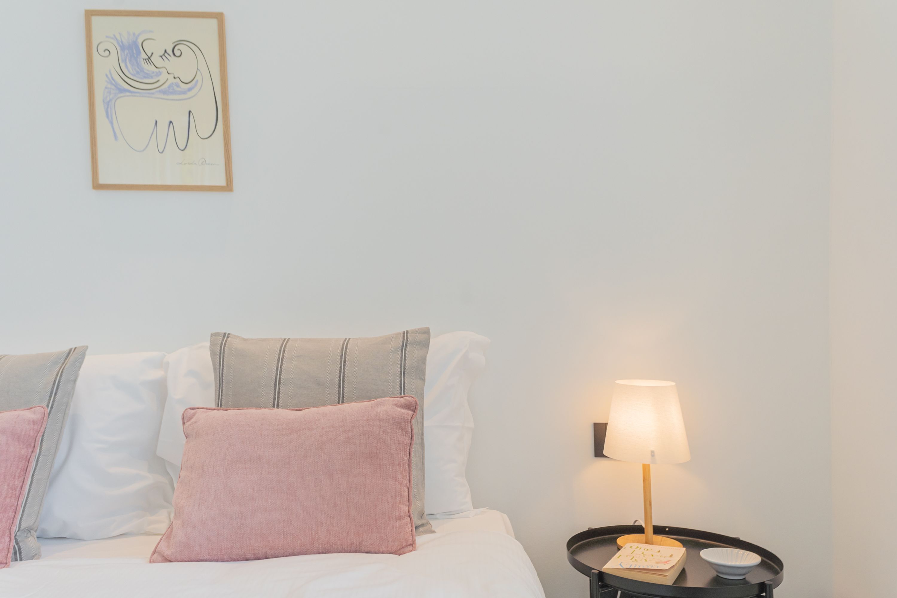 Una cama bien hecha con almohadas, una mesita de noche con lámpara y una obra enmarcada en la pared.