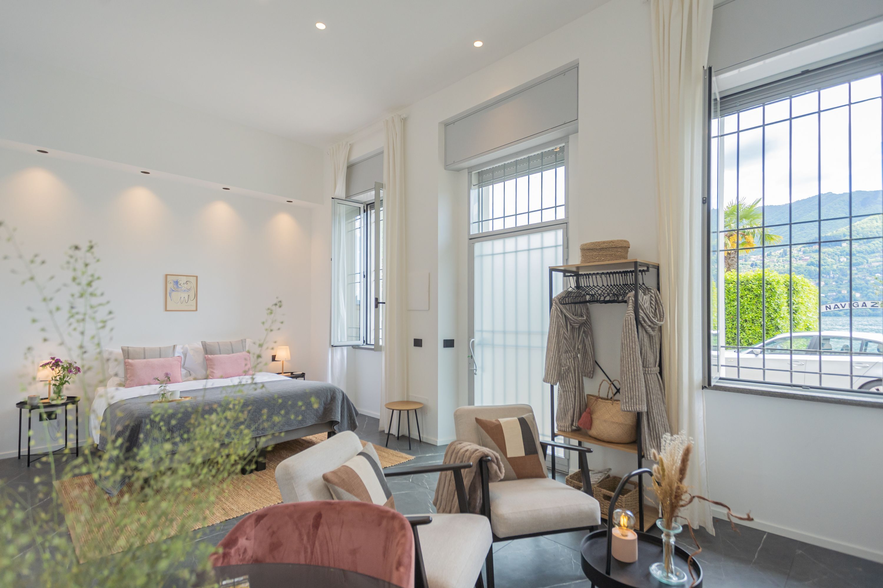 A modern bedroom with a large bed, a seating area with chairs, a small table, and a view of the outdoors.