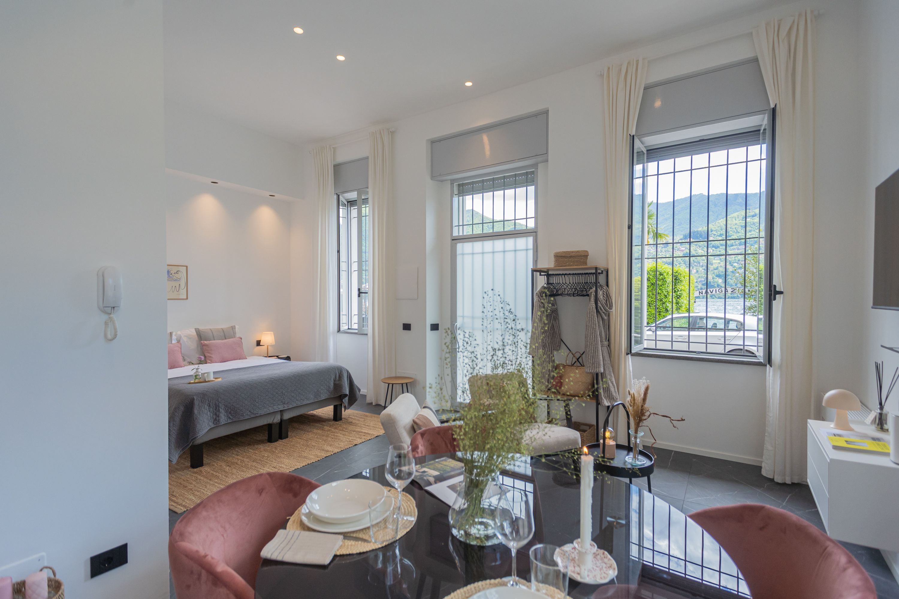 A modern hotel room with a bed, a dining table set for two, and a large window offering a view of the outdoors.