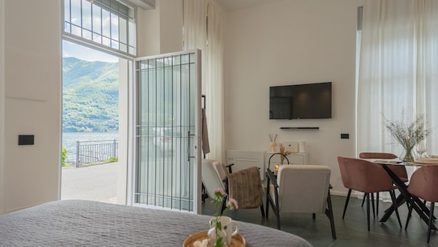 A hotel room with a large bed, a dining area with chairs, a TV mounted on the wall, and a view of a lake through a sliding door. - Piccola Como Lotus With 1 Bathrooms (Carate Urio)