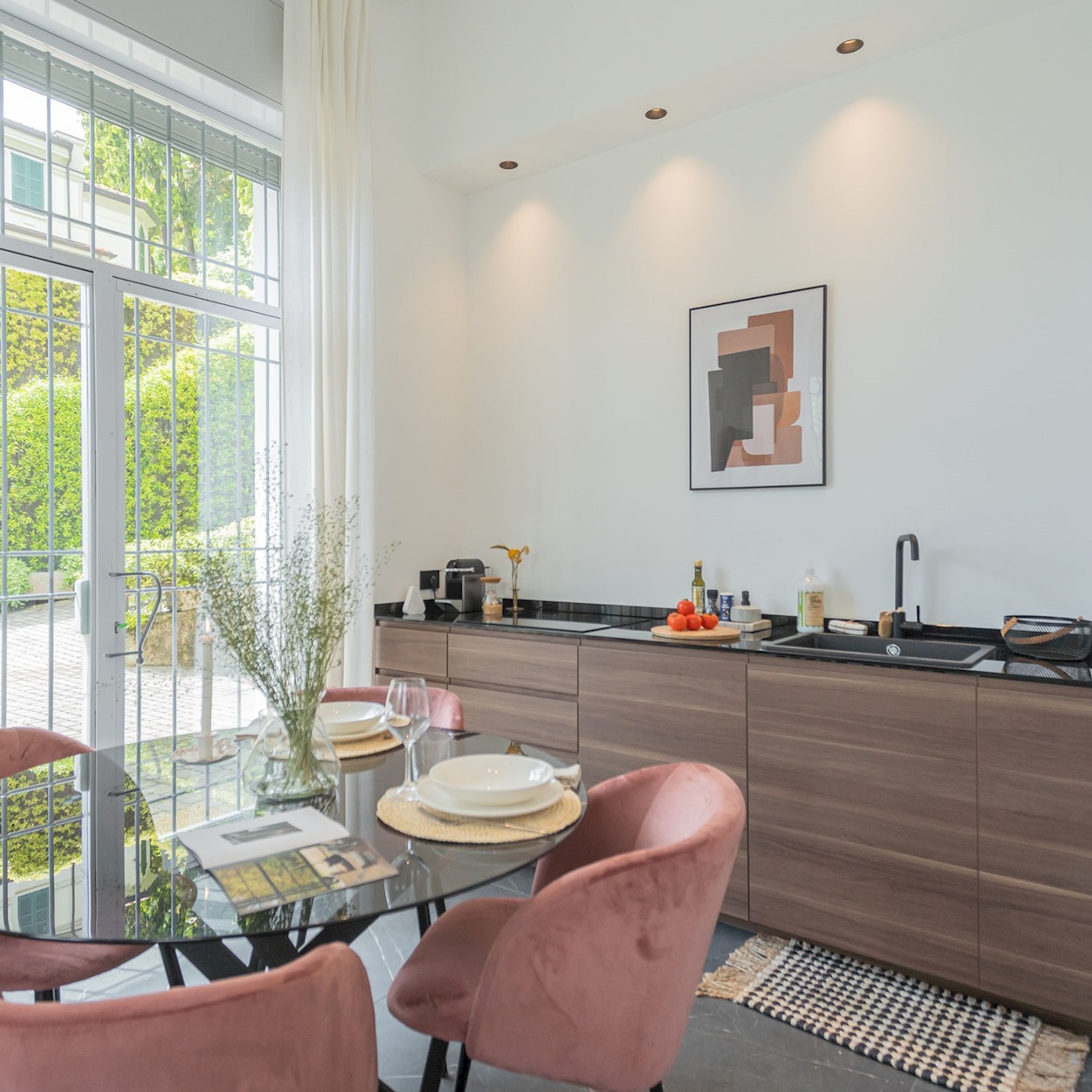 A modern kitchen with a glass dining table, pink chairs, and a view of greenery outside.