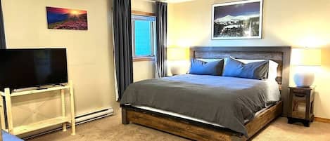Una habitación de hotel con cama, televisor sobre un mueble, ventana con cortinas y un cuadro en la pared.