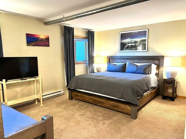 Una habitación de hotel con cama, televisor sobre un mueble, ventana con cortinas y un cuadro en la pared.