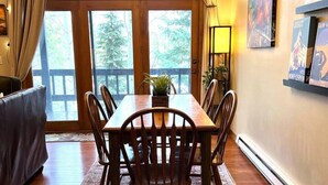 A dining area with a wooden table, chairs, a rug, and a view through large windows.