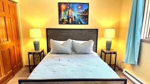 A bed with a wooden frame, two bedside tables with lamps, a painting above the bed, and a window with curtains.