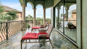 A covered balcony with wicker furniture and red cushions, large windows, and a view of a building with a tiled roof. - Monaco (Miramar Beach)